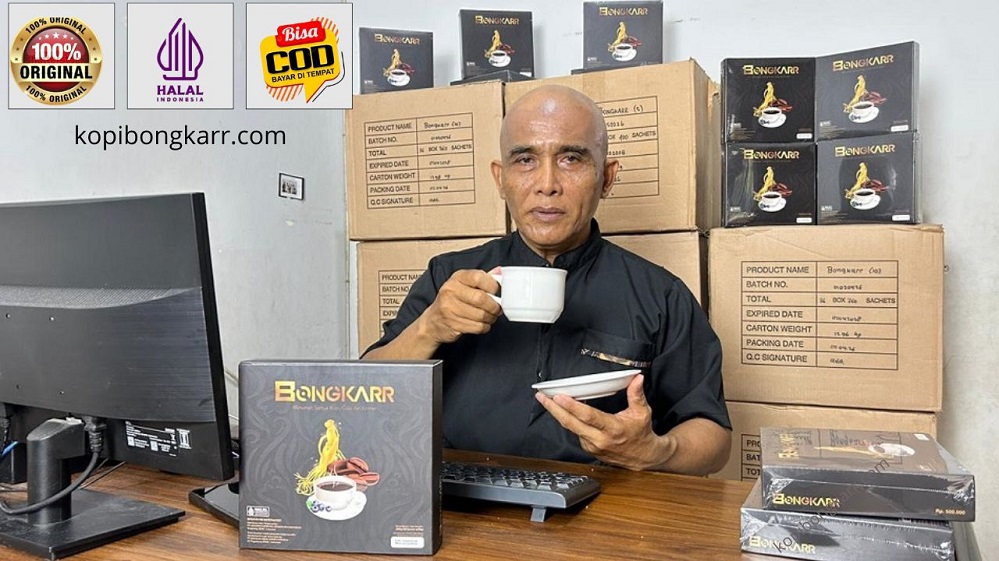Foto asli agen resmi sedang membuktikan sendiri khasiat kopi bongkar dengan menikmati secangkir kopi di meja kerja, berlatar kemasan box dan tumpukan kardus stok siap kirim.