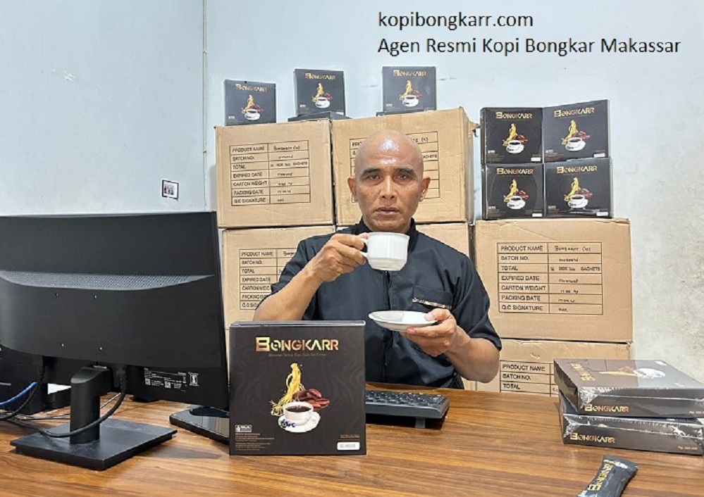 Penulis review Kopi Bongkar Makassar sedang menikmati secangkir kopi stamina di meja kerja, dengan latar belakang ketersediaan puluhan karton stok Kopi Bongkar original yang siap dikirim ke Sulawesi.