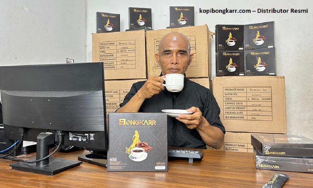 Agen dan distributor resmi Kopi Bongkar asli sedang menikmati secangkir kopi herbal di ruang kerja, dengan latar belakang ketersediaan stok puluhan karton produk original yang siap dikirim ke konsumen.